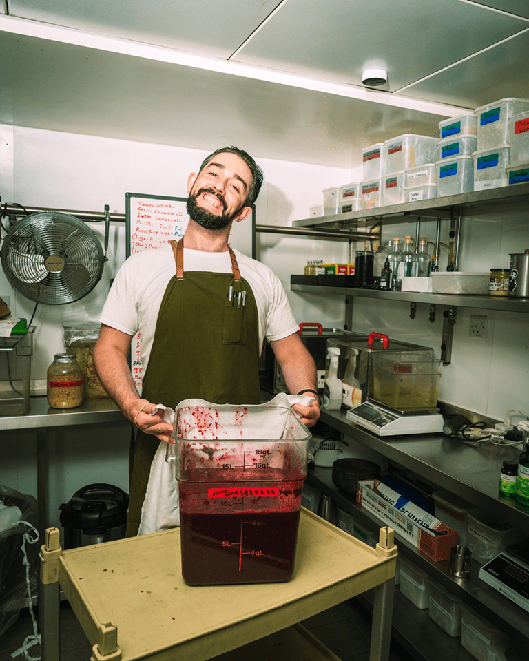 Person in an apron smiling, holding a large container of red liquid in a kitchen with shelves of labeled boxes and kitchen supplies.