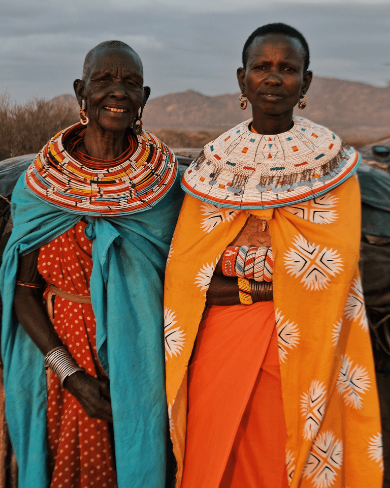 Two women in colorful traditional attire and beaded necklaces stand together outdoors, against a backdrop of hills and a cloudy sky.
