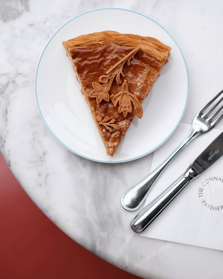 A slice of pie with decorative floral designs on a white plate, next to a silver fork and knife on a marble table.