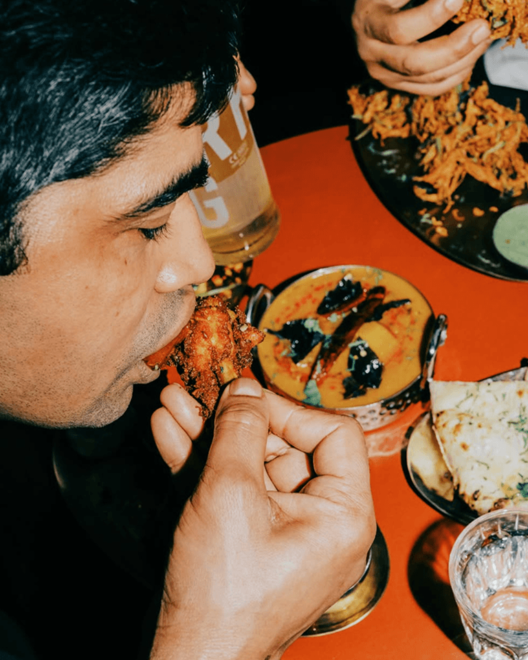 A person eating spicy fried chicken with various Indian dishes on a table, including naan bread, dal, and water in a glass.