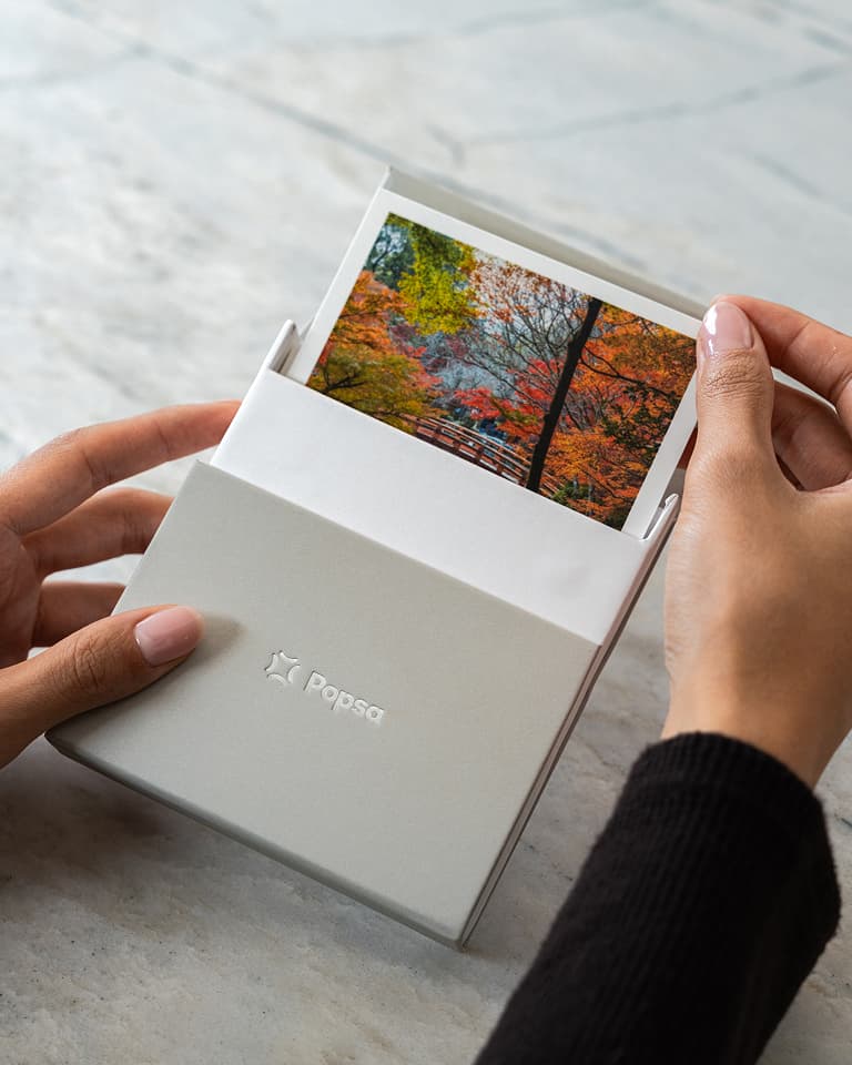 Hands holding a Popsa photo box, revealing a picture of colorful autumn trees.