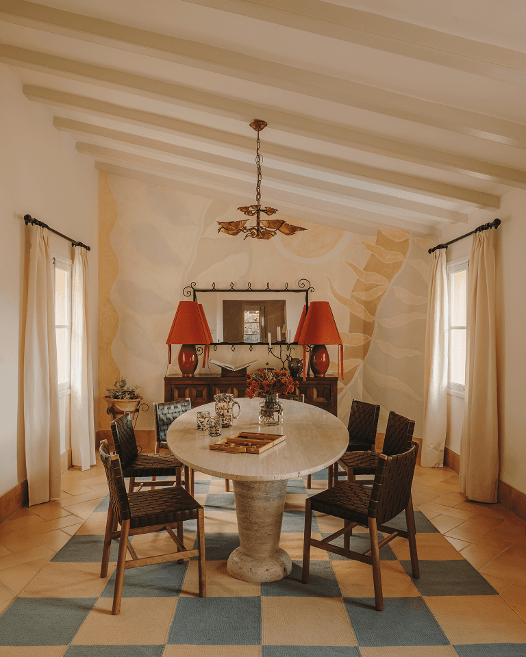 Cozy dining room with a round table, four wicker chairs, red lamps, mural wall art, and arched windows with curtains.