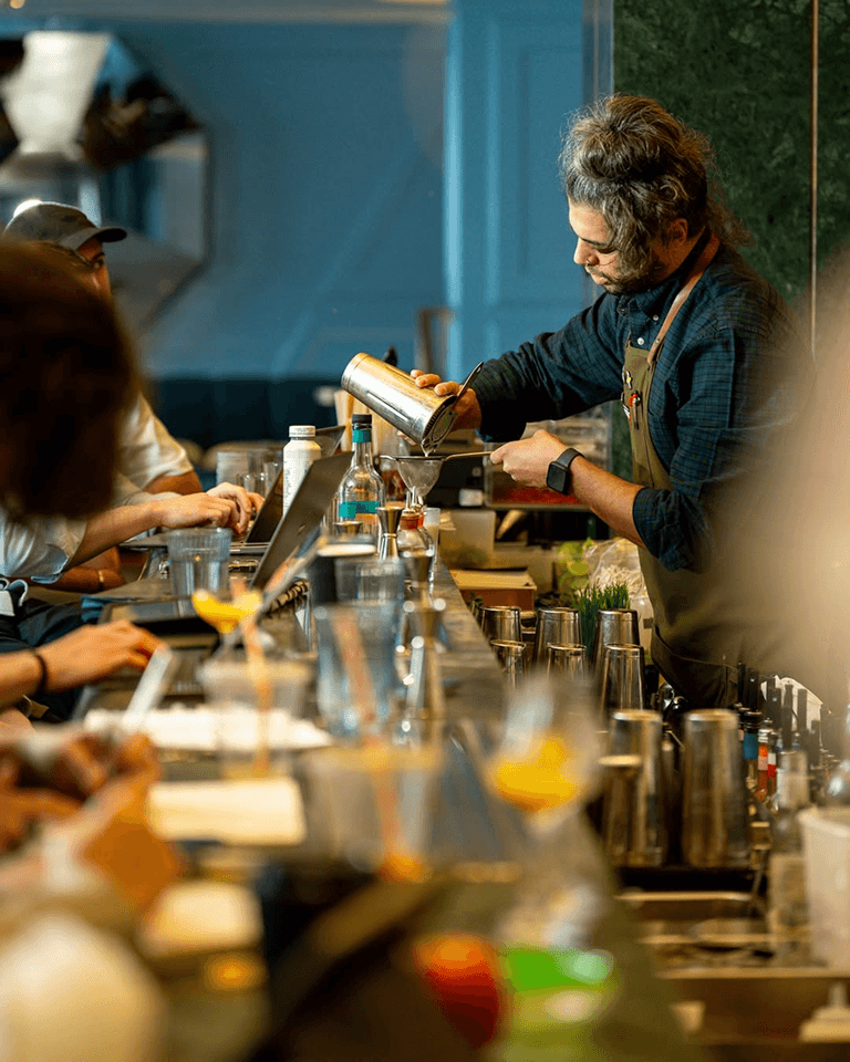 A bartender pours a drink from a shaker while people sit at the bar with laptops and drinks in a modern bar setting.