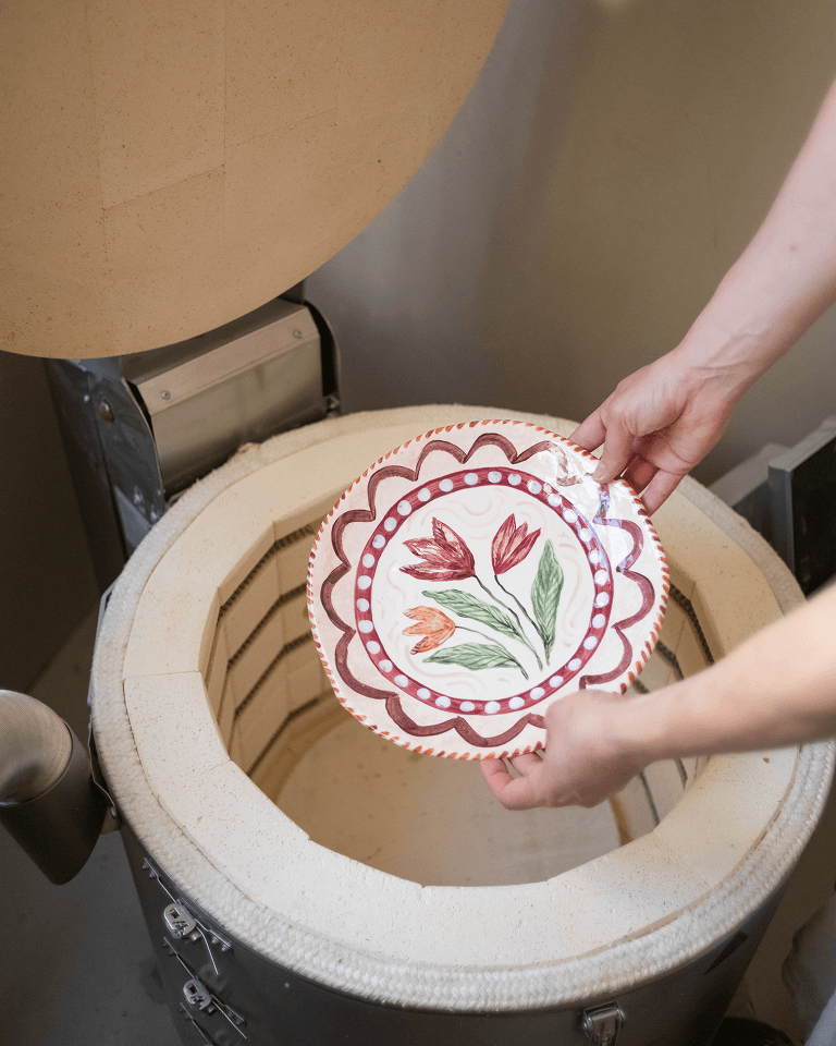 Hands placing a decorative plate with floral design into an open pottery kiln.