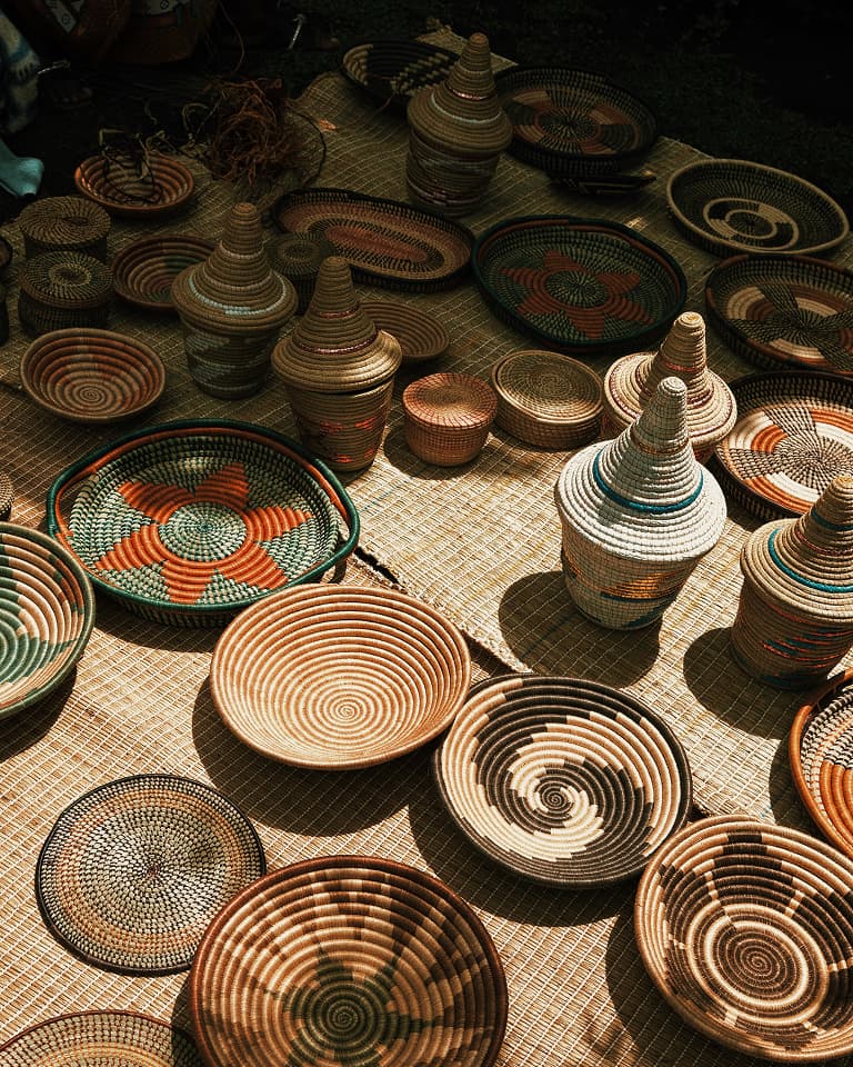 A variety of colorful woven baskets and lids with intricate patterns are displayed on a straw mat under sunlight.
