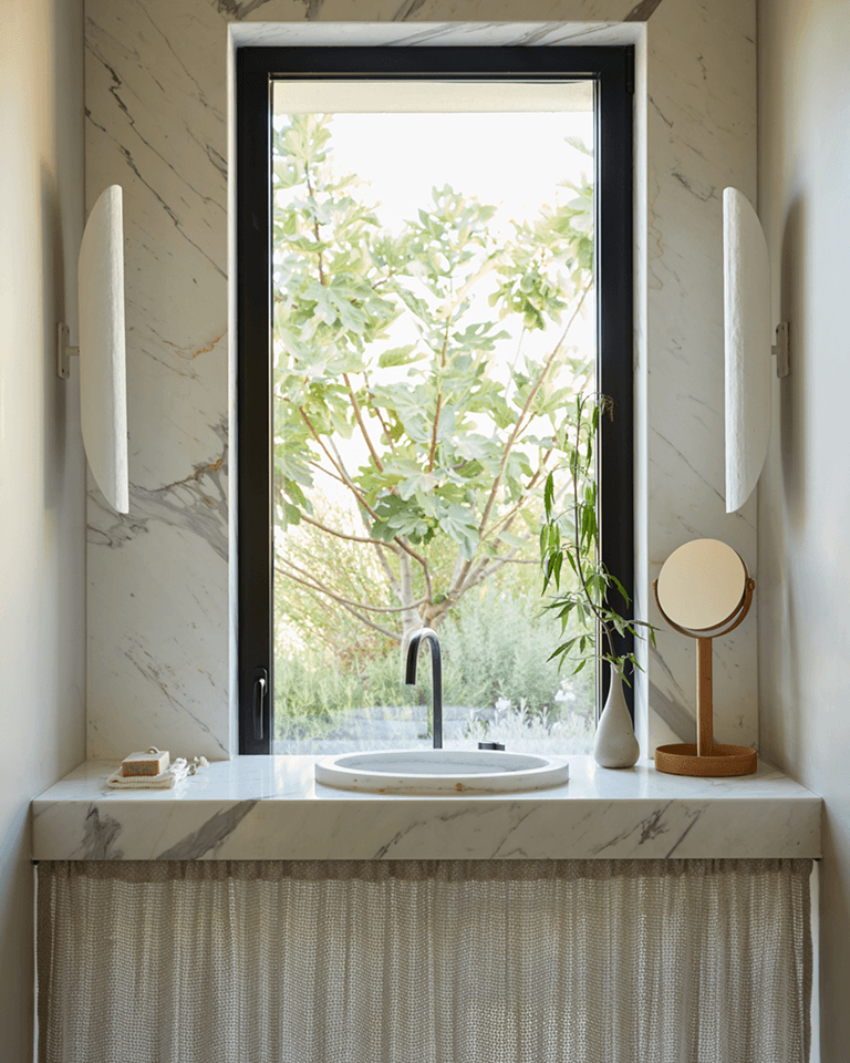 Marble sink area with a round mirror, vase, and soap dish, set in front of a large window showing a leafy tree outside.