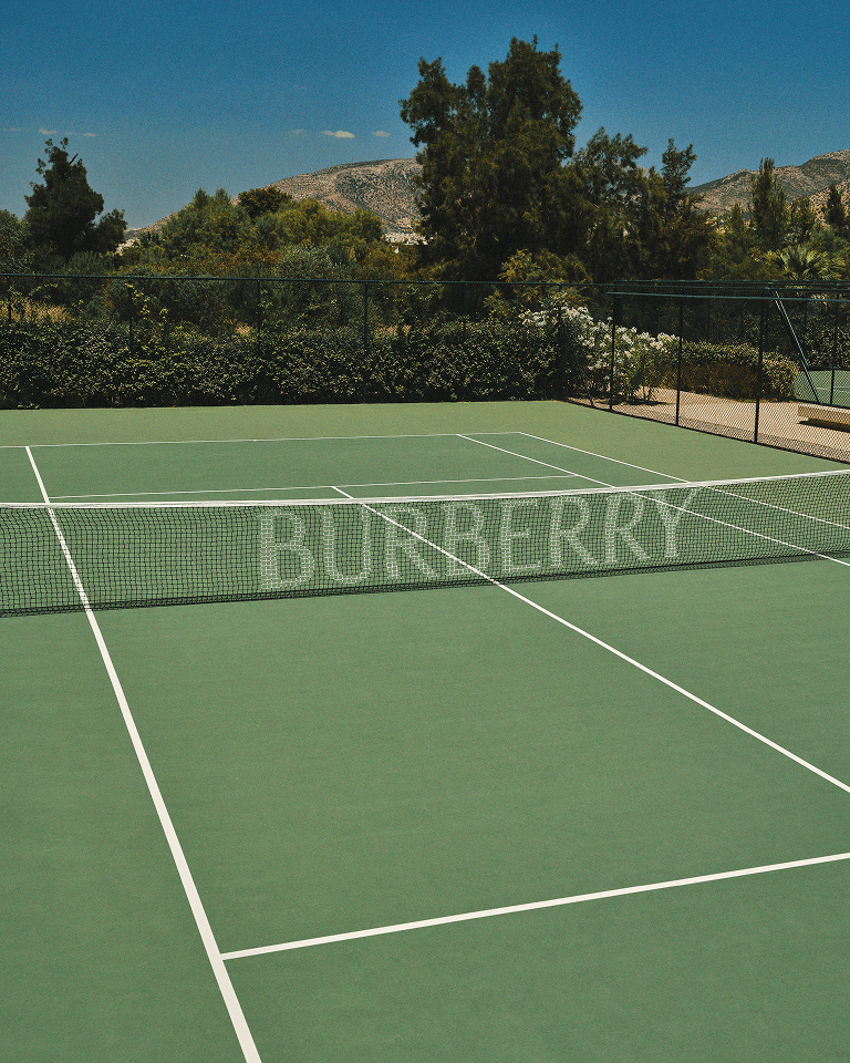 A green tennis court with a net and "Burberry" logo, surrounded by trees and mountains under a clear blue sky.