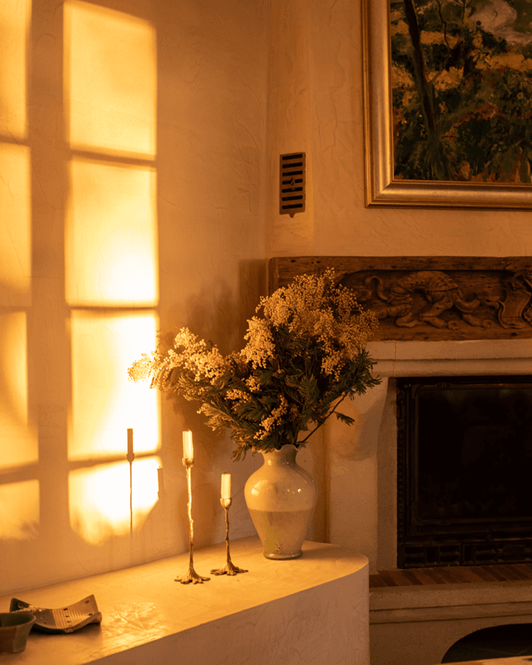 Sunlit living room corner with a vase of flowers on a mantel, three candlesticks, and a painting above a fireplace casting shadows on the wall.