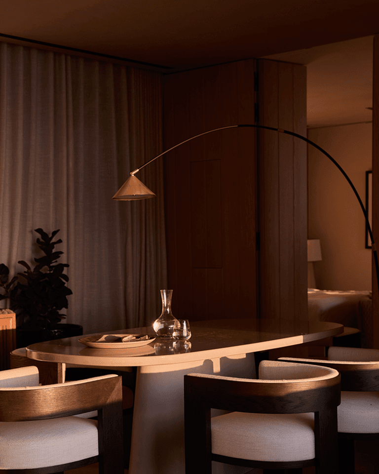 Cozy dimly-lit dining area with a round table, wooden chairs, arched floor lamp, curtains, and a plant in the background.