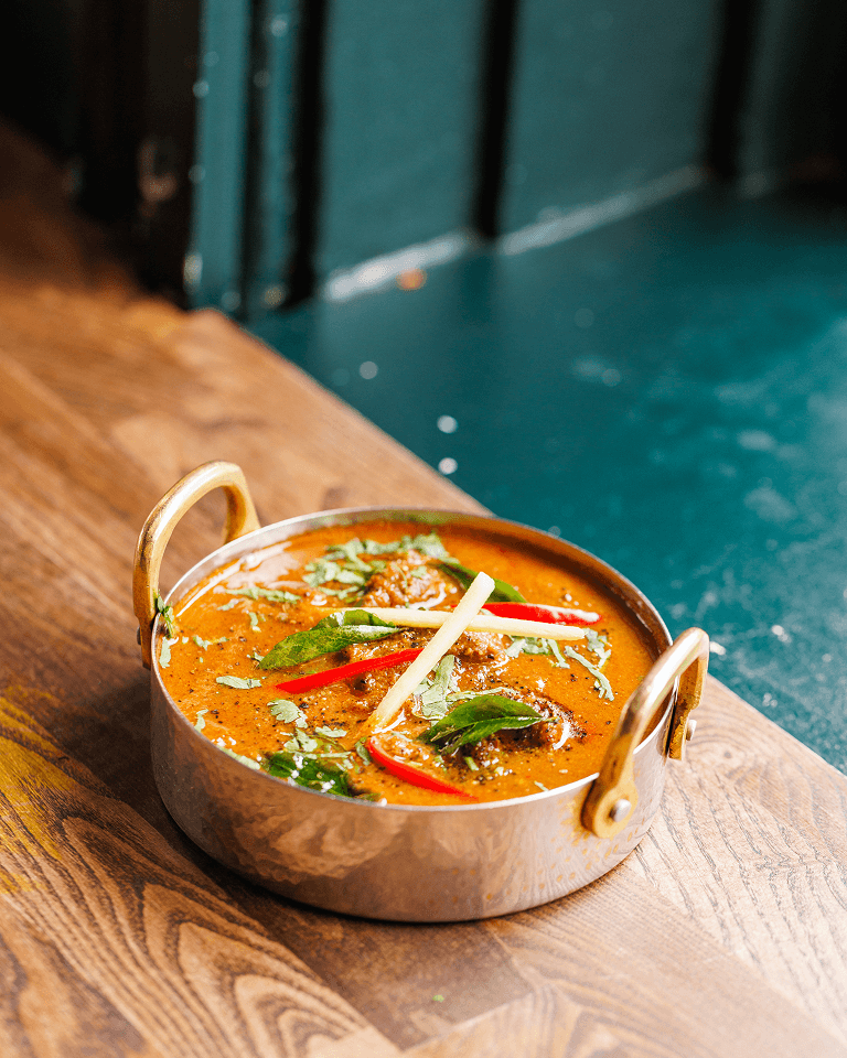 A bowl of spicy curry with red peppers, herbs, and sliced ginger served in a brass vessel on a wooden table.