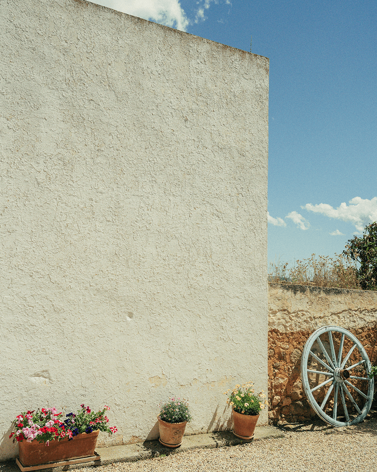 Weathered stucco wall with potted flowers at its base, clear blue sky above, and a rustic wagon wheel leaning against a short stone wall.
