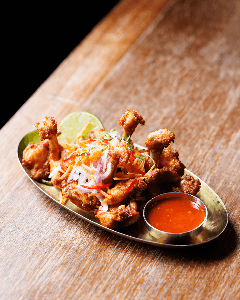 Fried chicken lollipops garnished with sliced onions and carrots, served with a lime wedge and a small bowl of red dipping sauce on a brass platter.