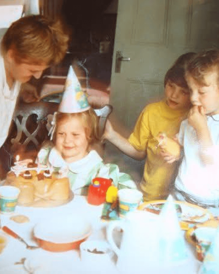 Children at a birthday party gather around a table with a cake, wearing party hats. One child appears excited as others look on.