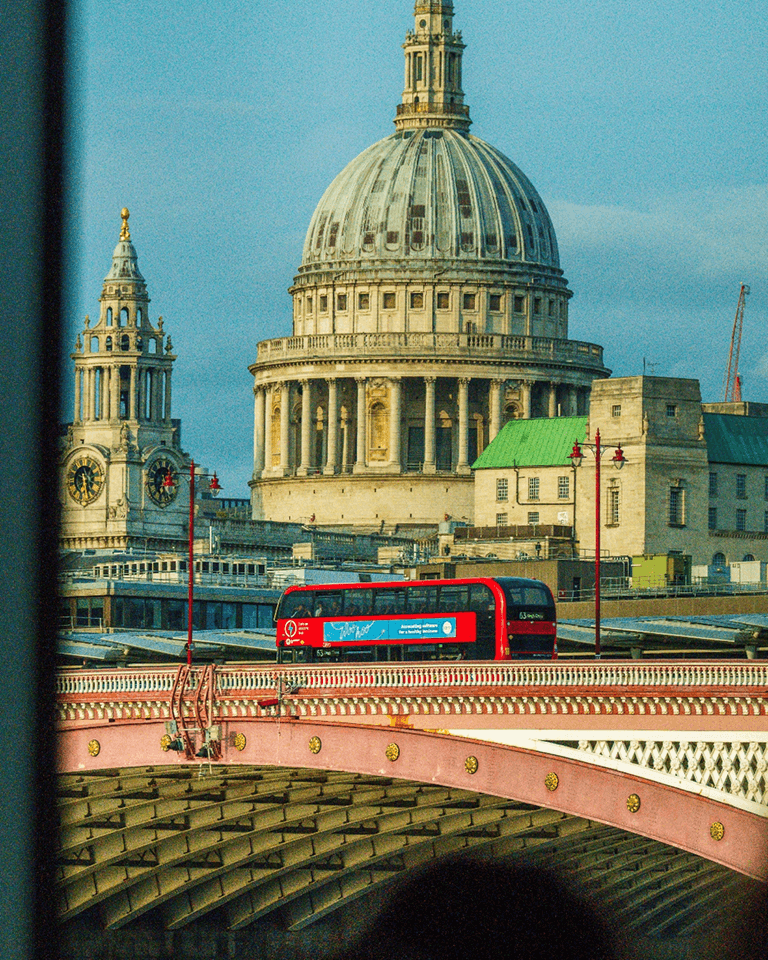 A red double-decker bus crosses a bridge in front of a large domed cathedral with a blue sky background.