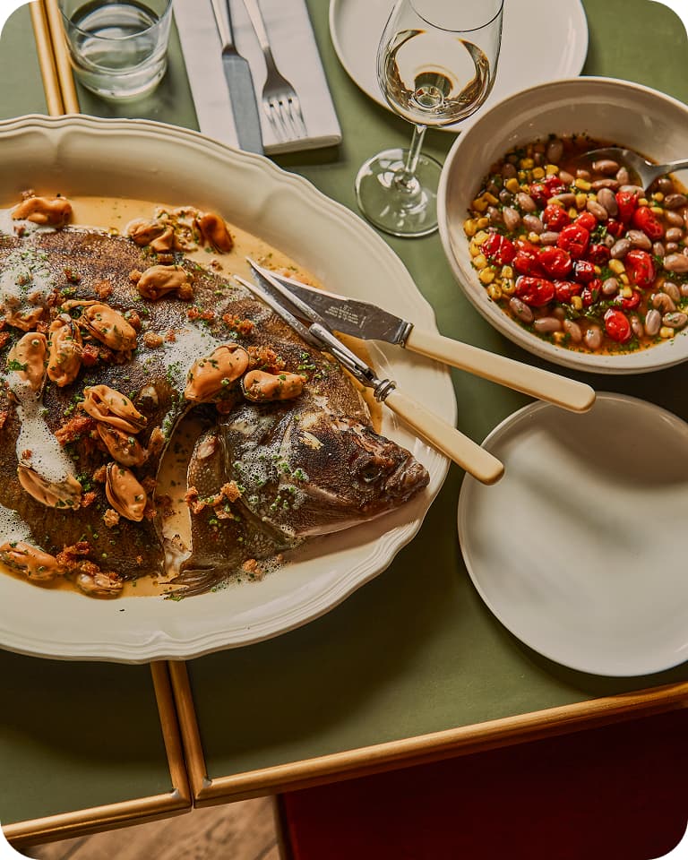 Whole roasted fish on a white platter topped with mussels and herbs, knives beside a bowl of bean and cherry tomato salad, wine glass and plates.
