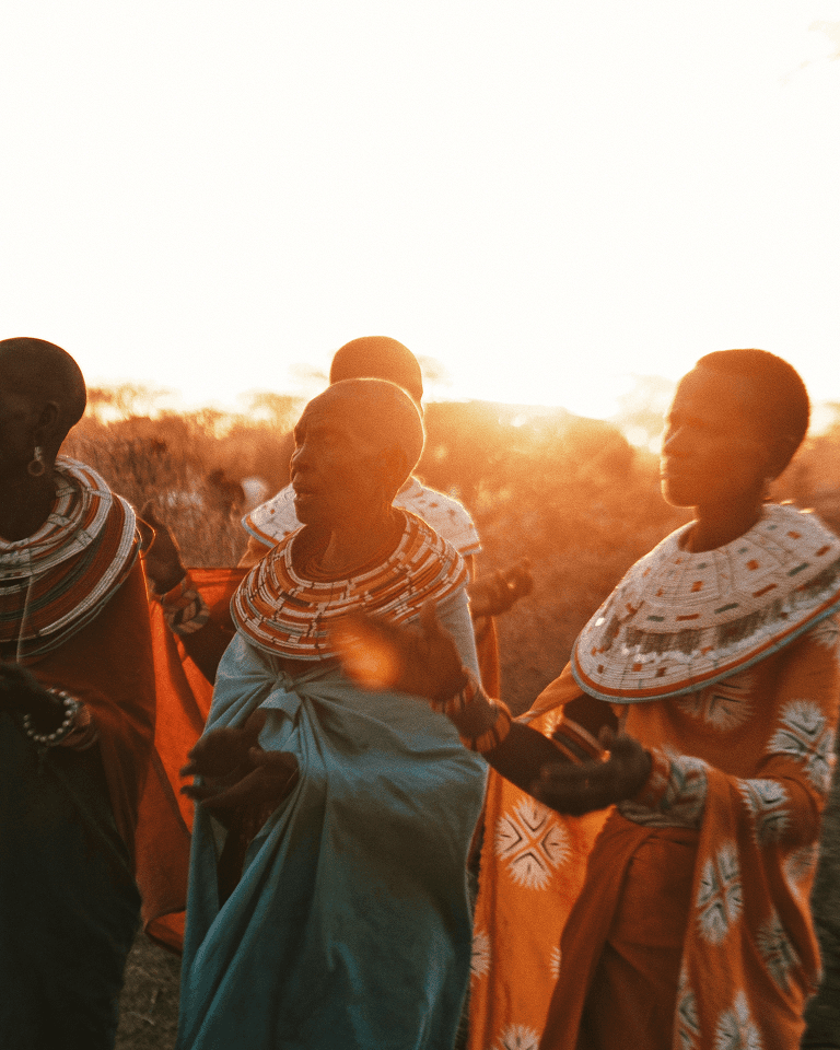 A group of people in colorful traditional attire, singing and clapping outdoors at sunset, with bright sunlight in the background.