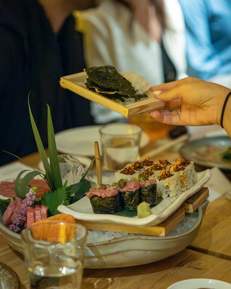 A hand holding a wooden plate with seaweed over a table of assorted sushi rolls, garnished with greens and flowers.