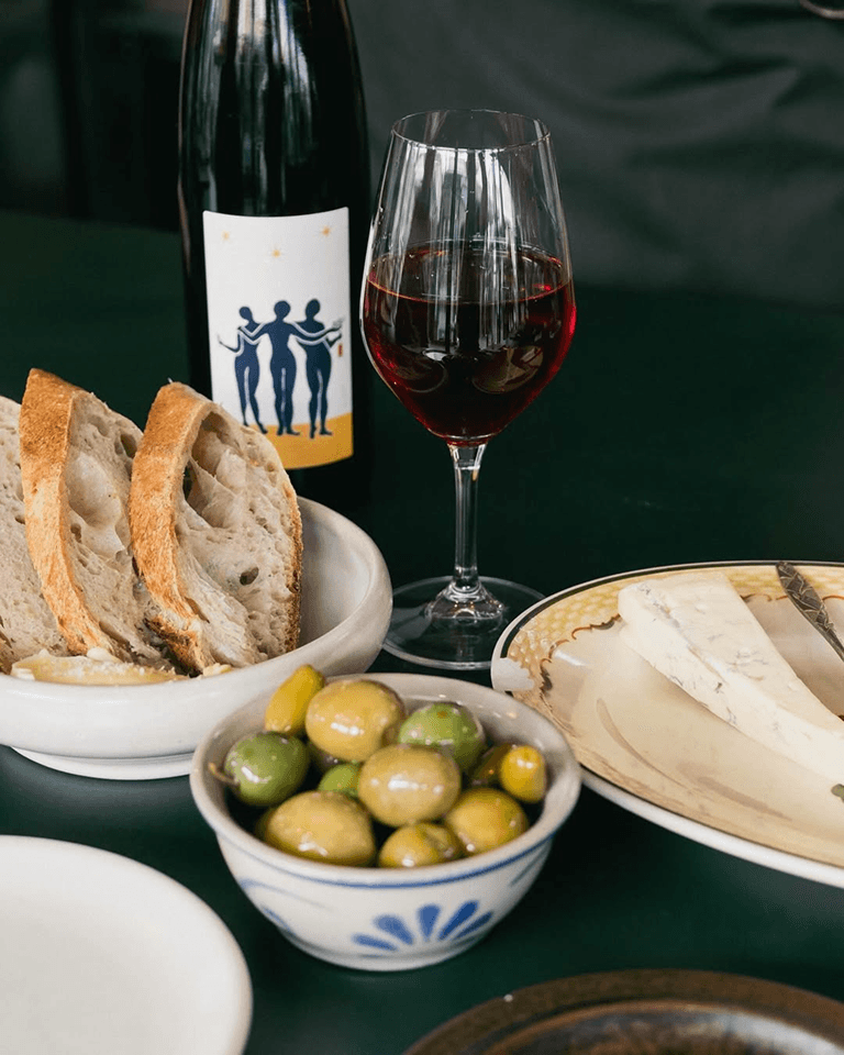 A bottle of red wine, a glass filled with wine, slices of bread, a bowl of green olives, and a plate of cheese are arranged on a dark green table.