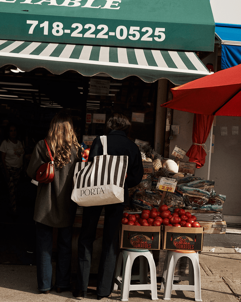 Two people stand outside a grocery store under a green-striped awning, with fruit and vegetable displays, including tomatoes, under a red umbrella.