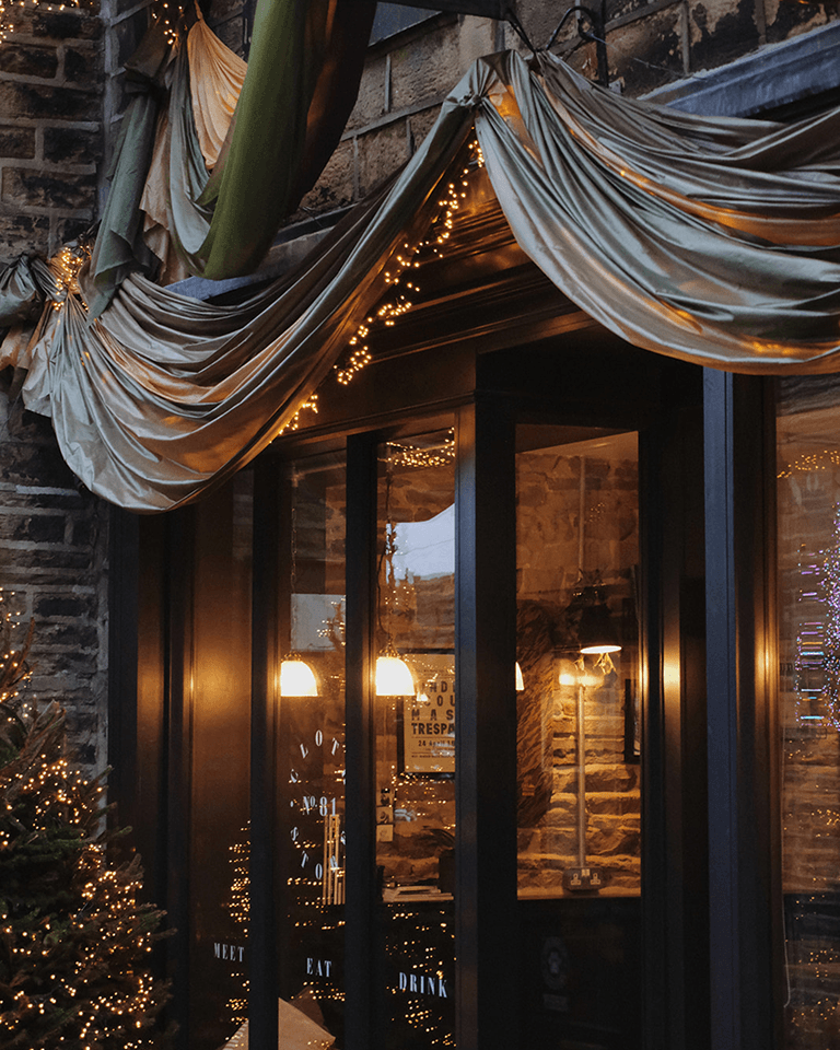 Cozy entrance of a stone building decorated with draped fabric and string lights, illuminated by warm interior lighting.