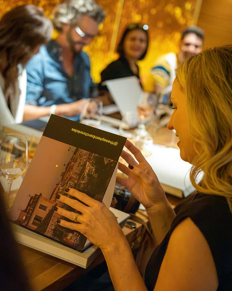 A woman at a dinner table examines a book with others in the background, creating a warm, social atmosphere.