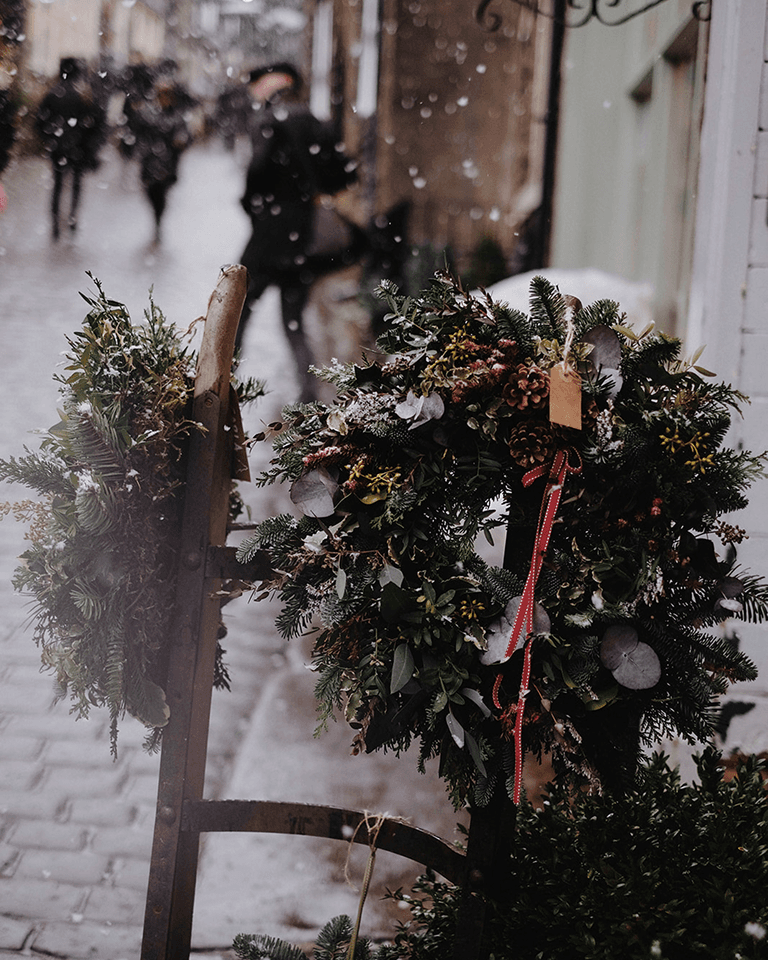 Snow falls on a cobblestone street adorned with festive wreaths, while blurred pedestrians walk in the background.