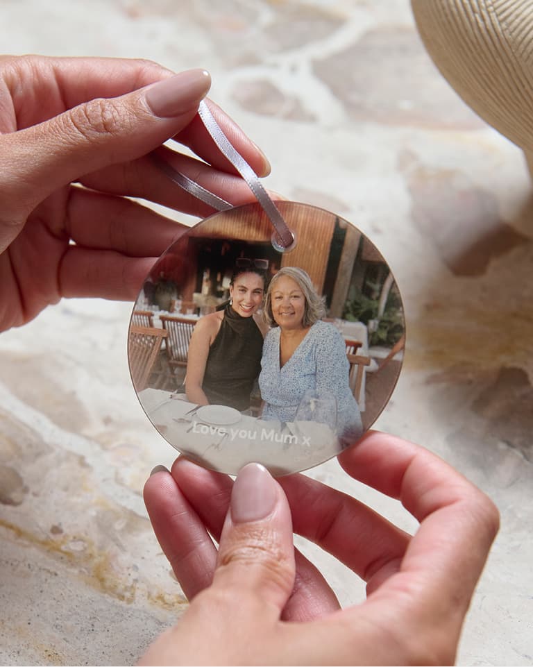Hands holding a circular photo ornament showing two smiling people seated outdoors, with "Love you Mum x" written beneath the image.