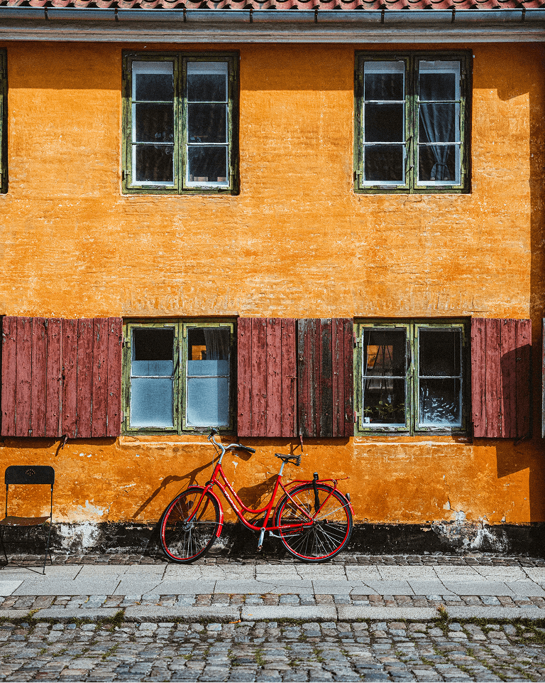 Red bicycle leaning against an orange wall with green-framed windows and red shutters, beside a black chair on a cobblestone street.