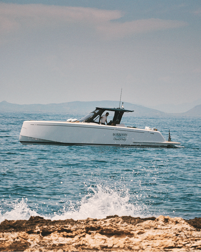 White boat with "BURBERRY" logo on the side, cruising on a calm sea with distant hills under a clear sky. Waves gently hit the rocky shore.