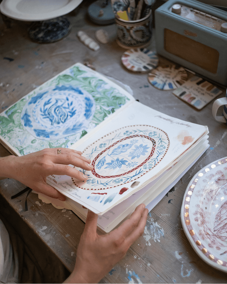 An open sketchbook with hand-painted floral designs on a cluttered wooden table, surrounded by pottery and a retro radio.