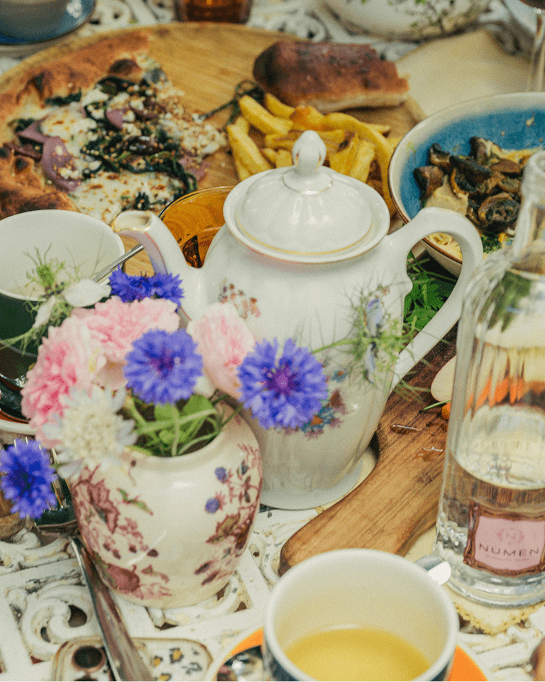 A floral teapot and vase beside a cup of tea, with pizza, fries, and other dishes on a patterned table.