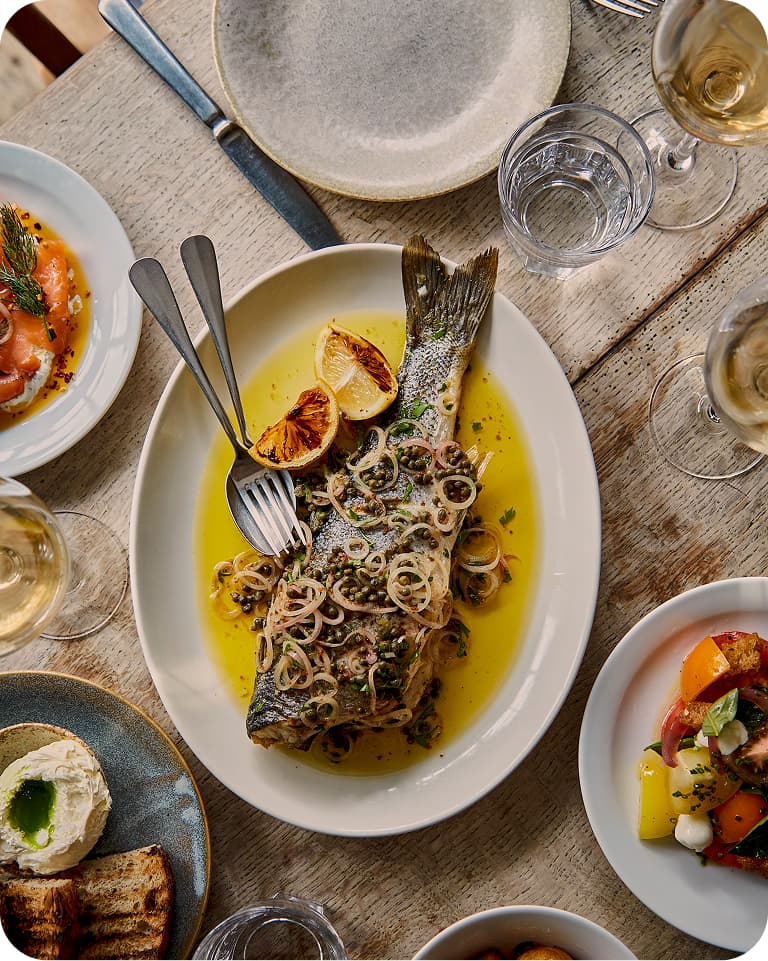 Whole grilled fish on a white platter with charred lemon halves, capers and shallots in olive oil, surrounded by plates, bread and wine glasses.