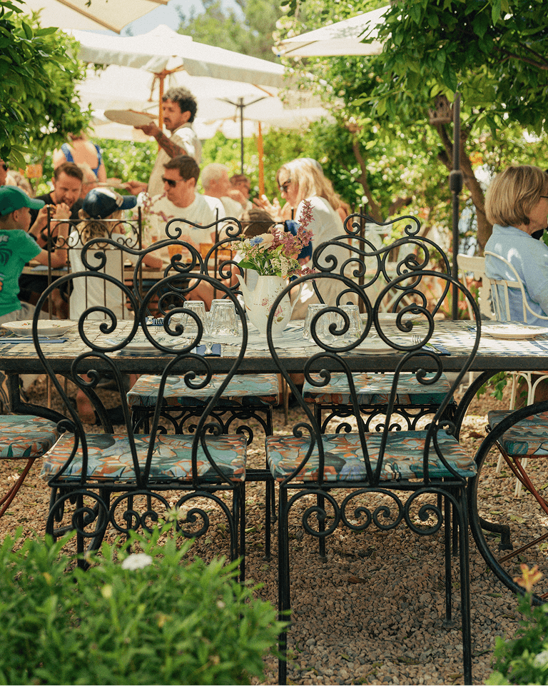 Ornate metal chairs around a table with a vase of flowers, set in a lush outdoor garden patio with people dining under umbrellas.