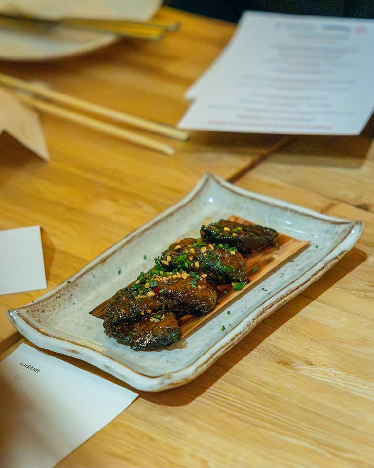 Steak slices garnished with chopped herbs and nuts on a rectangular ceramic plate, placed on a wooden table with menus and chopsticks nearby.