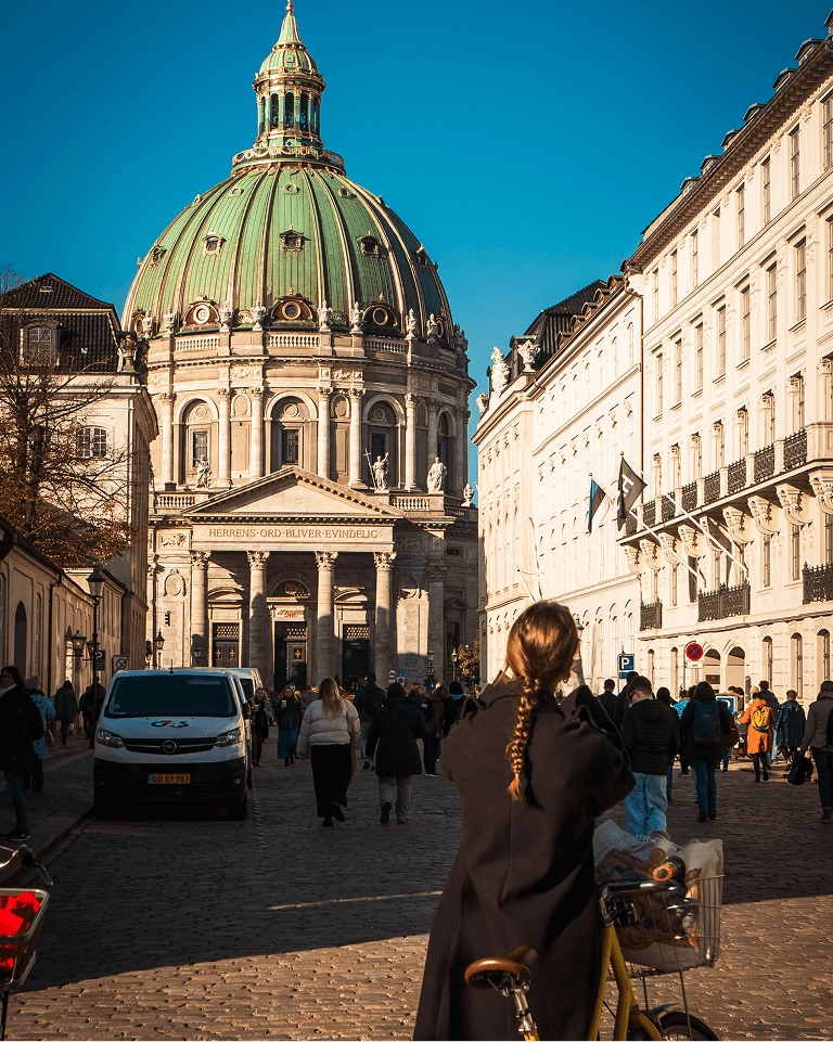 Woman photographing a historic building with a green dome surrounded by people and city architecture in bright sunlight.