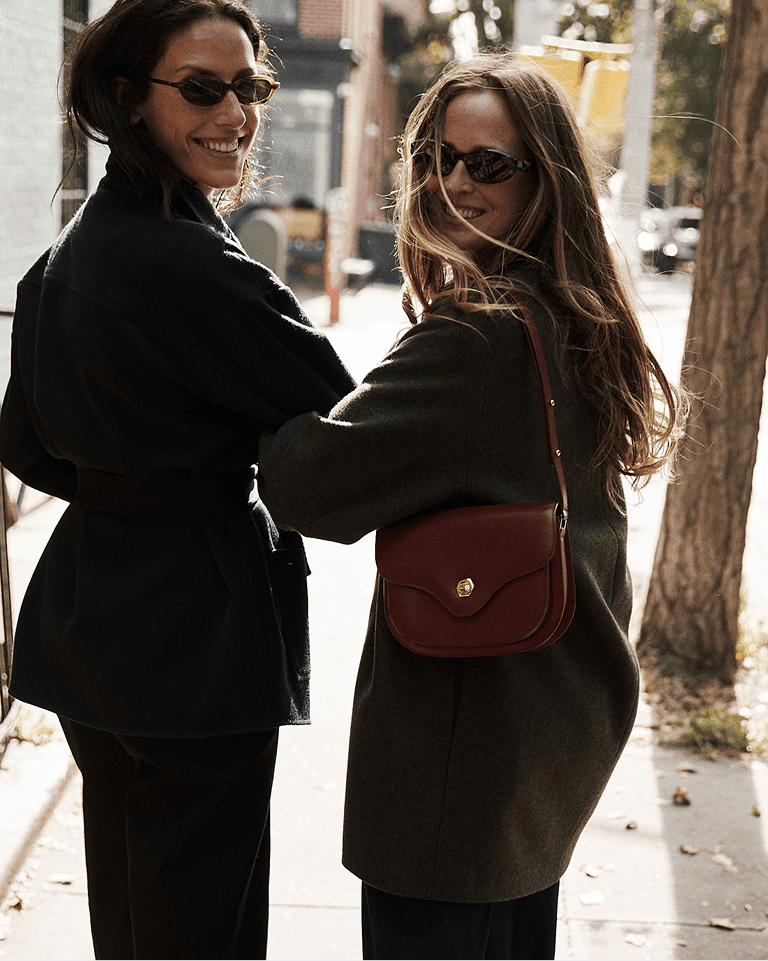 Two women, wearing sunglasses and dark coats, smile while standing on a sunlit street, one carrying a red shoulder bag.