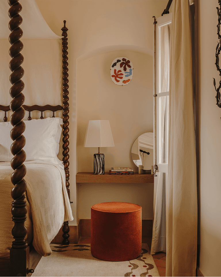 Cozy bedroom corner with a wooden four-poster bed, a small side table, a lamp, books, a mirror, and a red ottoman under a decorative wall piece.