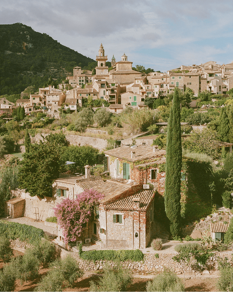 A picturesque hillside village with rustic stone houses, lush greenery, and a tall cypress tree, under a partly cloudy sky.