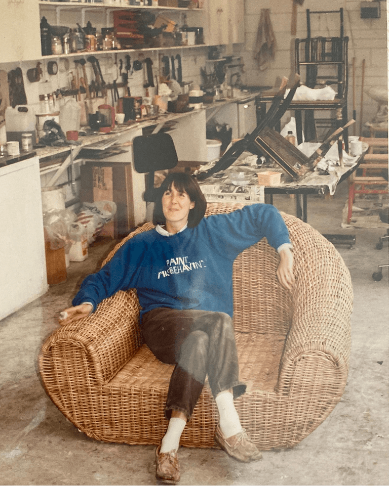 A person in a blue sweater sits relaxed on a wicker chair in a cluttered art studio with shelves, art supplies, and tools in the background.