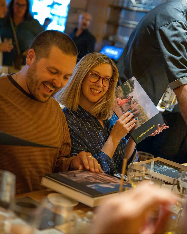 A smiling man and woman sitting together at a table, each holding a book, with drinks and blurred people in the background.