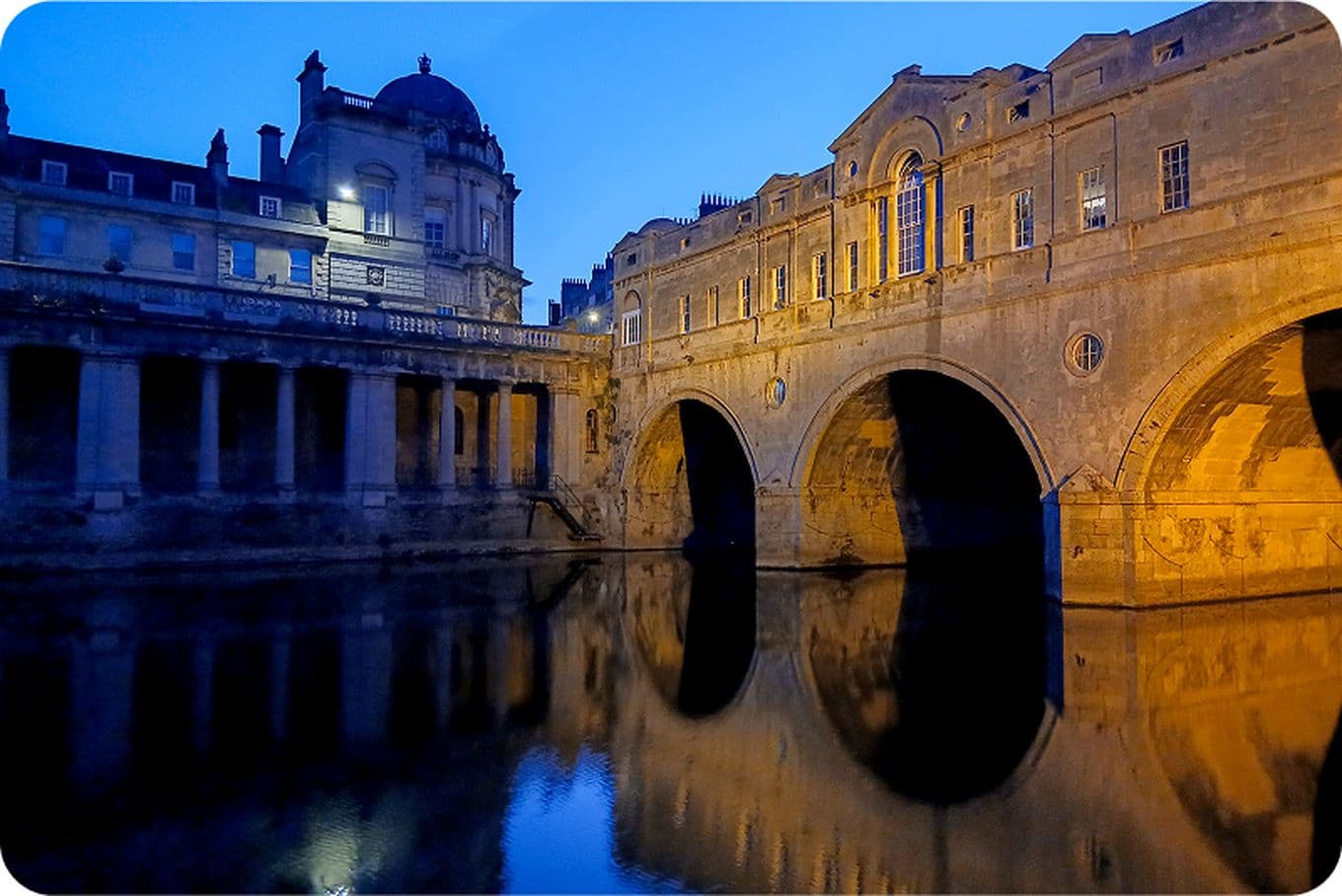Historic stone bridge and buildings reflected in a calm river at dusk, with a deep blue sky and warm yellow lighting.