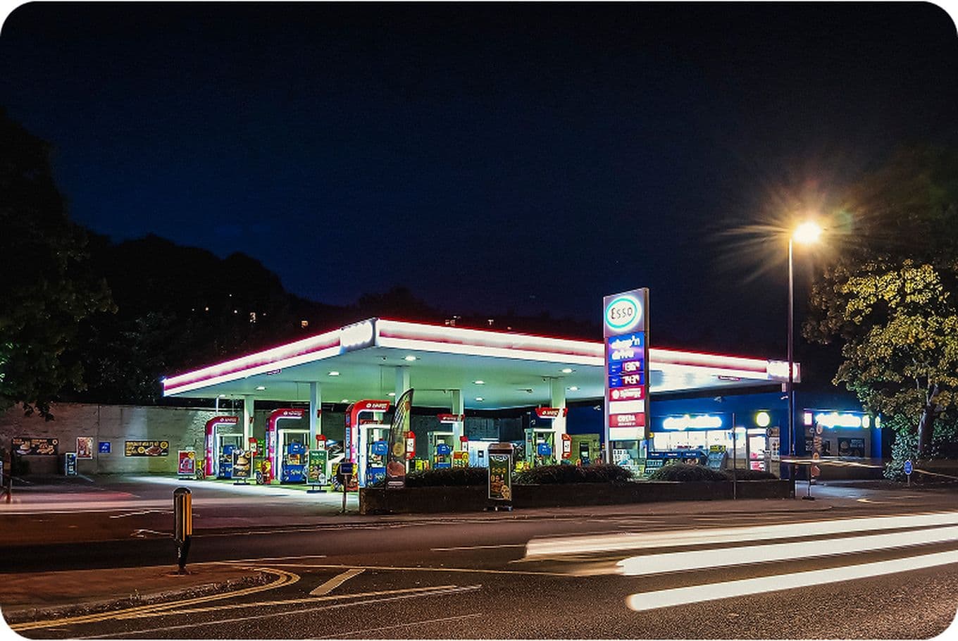 Night view of a brightly lit gas station with multiple fuel pumps, a glowing sign displaying prices, and light trails from passing vehicles.