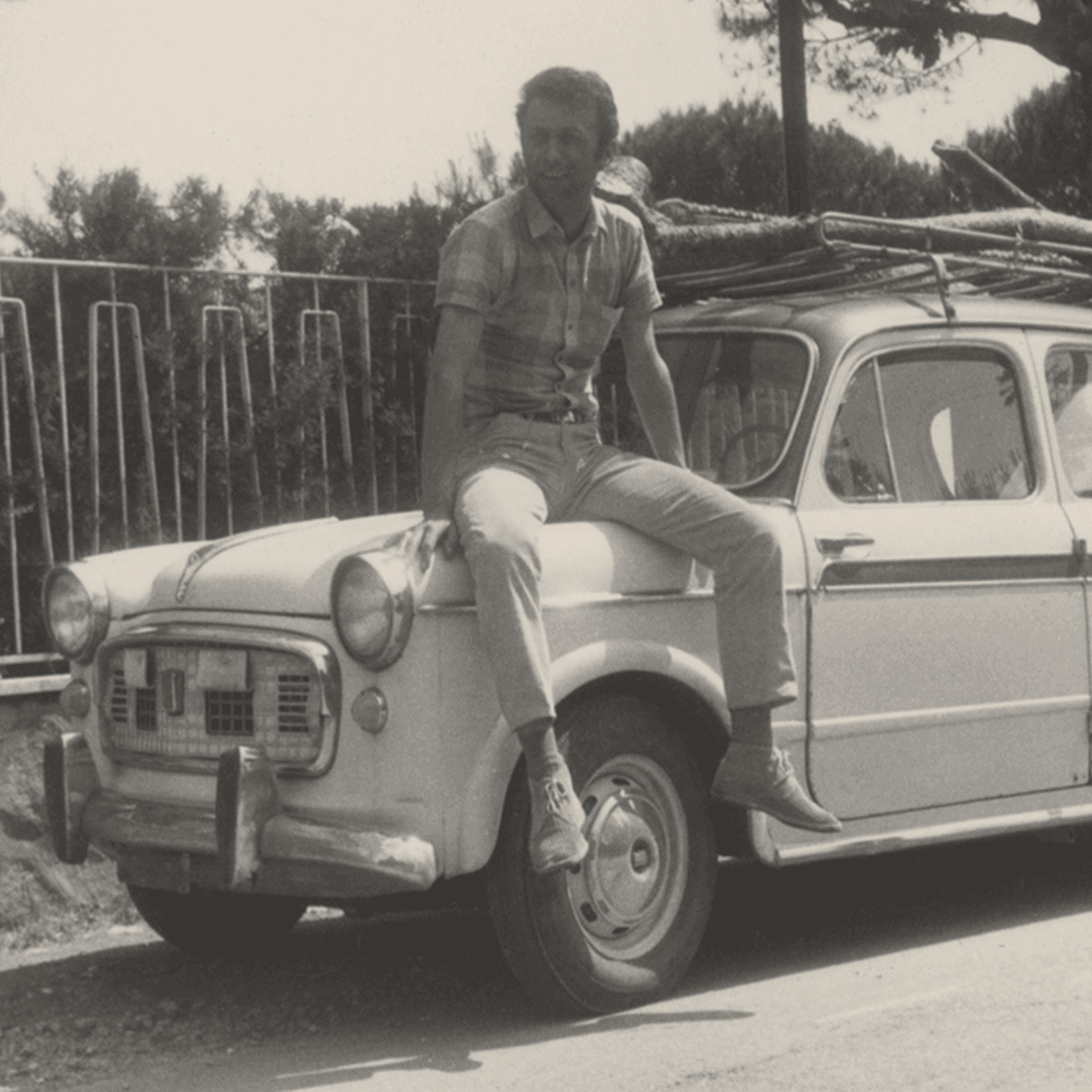 Man sitting on the bonnet of a vintage car, wearing a short-sleeve shirt and trousers/
