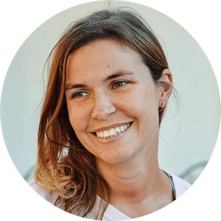 Smiling woman with long brown hair, wearing a light-colored top, looking slightly to the side against a plain background.