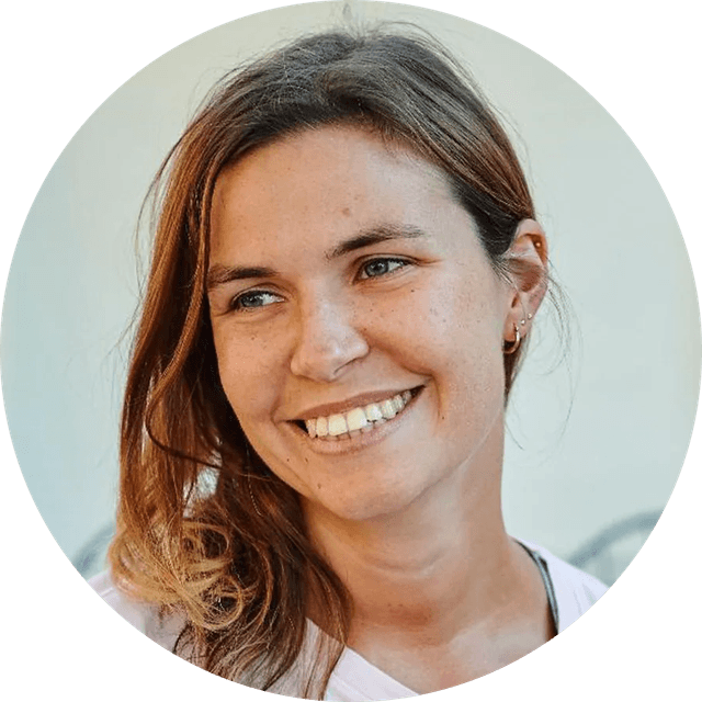 Smiling woman with long brown hair, wearing a light-colored top, looking slightly to the side against a plain background.